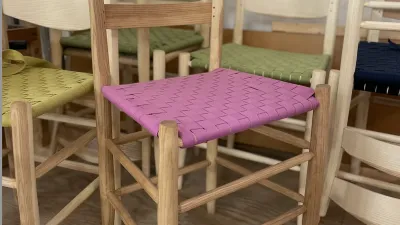 Several ladder back chairs are shown, some stacked on each other, in a variety of states of finish. There is a chair in the foreground with a pink wovven 