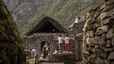 In the foreground, old stone walls frame the sides of the photo. Between and behind them, people can be seen working on a stone house in a lush, mountainous region. There are forested hills visible in the far background.