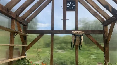 Interior shot of a timber frame greenhouse. Farm is visible outside of the clear walls and roof, and there are plants inside.