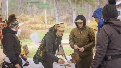 Several people standing outside look at a piece of lumber. The teacher is pointing at the lumber to show the students measurements.