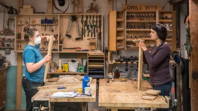 Two women at work benches facing each other measure pieces of wood in the woodshop