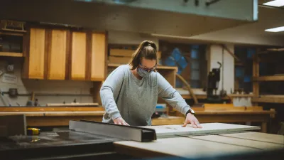 A person wearing a grey shirt and hair in a ponytail makes a cut on the table saw.