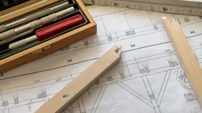 A close up of a drafting table with writing implements, building plans and wooden model making material. 
