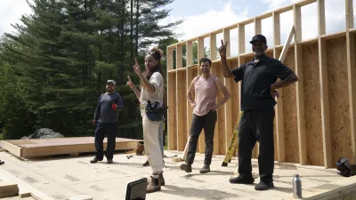 Four people are standing on a floor deck with one framed wall raised behind them. The four people are evenly spaced and facing the camera. Some are smiling and two are holding up peace signs.