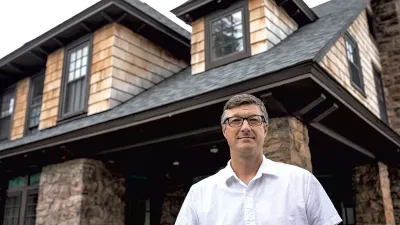 Devin Coleman stands in front of a cottage with stone and wood siding and gables. Devin is wearing a short sleeved white button down shirt, glasses, and short grey hair.