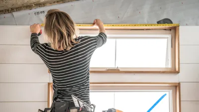 A person with blonde hair and black-and-white striped shirt uses a measuring tape on the top of a door frame