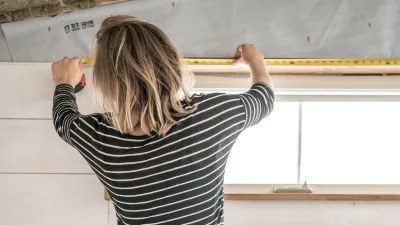 A person shot from behind wearing a black and white striped shirt and toolbelt uses a tape measure on the top of a doorframe.
