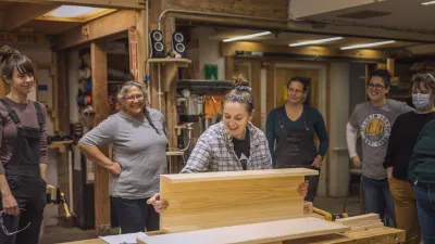 An instructor holds a piece of wood on a work bench, demonstrating for students standing behind.