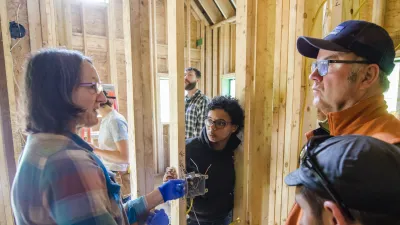 Students listen to an instructor gathered around a roughed-in electrical switch in a framed building.
