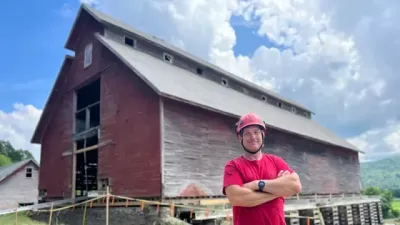 Eliot Lothrup stands in from of the East Monitor Barn, wearing a hard hat and red t-shirt, standing with his arms crossed. It is a sunny day.