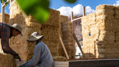 Two people crouch near a strawbale in front of a partially framed building.