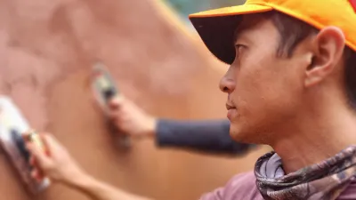 A person wearing an orange cap and neck gaiter carefully applies a smooth layer of reddish-brown clay plaster to a wall using a trowel. Another person’s hand is visible in the background, also plastering.