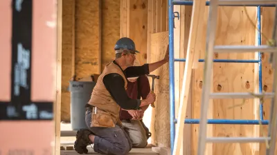 A person uses a hammer while kneeling inside a framed building with a hard hat on