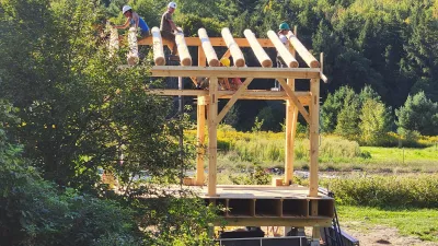 A group of people wearing safety helmets work together to construct a timber frame structure using round logs in a scenic outdoor setting. The partially built frame stands on a wooden platform surrounded by green grass, trees, and rolling forested hills under a clear blue sky.
