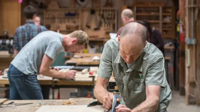 Two people, one in the foreground and one in the background, use hand planers in the woodshop.