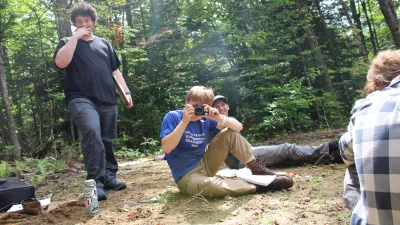 Three semester students are pictured in a sunny wooded clearing. The person in the center is pointing a camera at the photo taker, and there is another person standing to their left and one sitting to their right with their back turned.