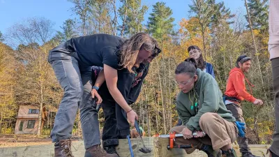 Two students toe-nail a floor frame joint together, with blue sky and fall leaves in the back.
