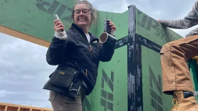 Sophia stands on a ladder in front of a zip-sheathed wall, holding a nailer and smiling at the camera