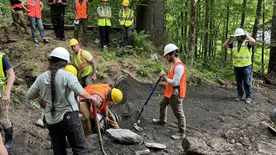 Gemini said A group of workers and students wearing safety vests and hard hats collaborate on a culvert installation project, using hand tools to move large stones and dark soil in a lush, wooded environment.