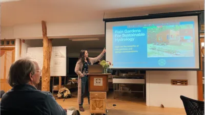 A speaker gestures at a presentation slide on a screen in a classroom.
