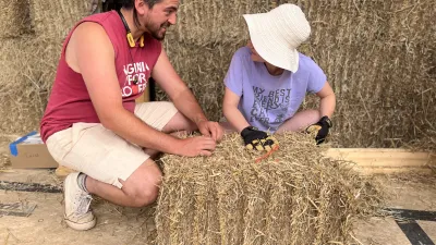 Two people work together shaping a straw bale inside a partially built structure made of straw walls. One person wears a sleeveless red shirt and light shorts, while the other wears a wide-brimmed hat, gloves, and a purple shirt.