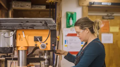 A student with blonde hair in a ponytail wearing a turquoise shirt uses the drill press in the woodshop.