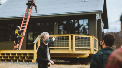 A man in the foreground gestures at a house in the background. Workers are installing solar panels on the roof.