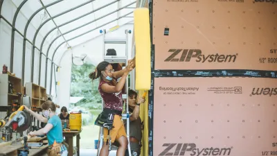 Students work on a tiny house in an outdoor classroom. A student with braids uses a nail gun on the tiny house siding in the foreground.