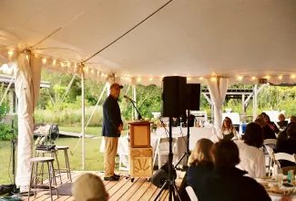 John Connell stands at a podium under a white celebration tent addressing a seated crowd. He is wearing a baseball cap and suit jacket.