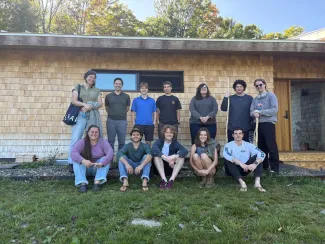The 2026 Design/Build semester poses for a group photo outside in front of a shingled cabin. There are two rows of students, some sitting down in front. It is a sunny day with blue skies.