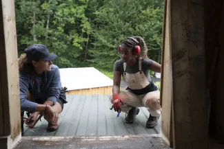 Two people crouch on a roof, seen through a doorway. They are both wearing ear protection, and both have curly brown hair. The person on the right is asking the person on the left a question.