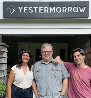 Three people stand smiling in front of the Yestermorrow front entrance sign. Left to right they are a shorter, dark haired femme person with a beige shirt and black pants, a man with a blue-gray light blue button down and glasses, and a man with longer, dark hair wearing a pink yestermorrow t-shirt