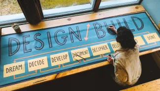 A photo from above of a dark haired woman painting a banner that says "Design/Build" with the steps of the design/build process below