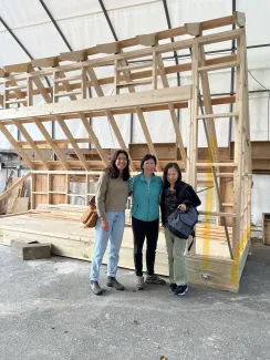 Three women of varying ages stand in front of a framed structure in an outdoor tent