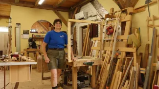Sally, a middle aged woman with short brown hair, blue t-shirt, and green cargo shorts, smiles at the camera in her shop. There are window and door frames, as well as woodworking tools and materials, all around.