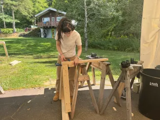 A semester student with long dark hair measures a piece of lumber on sawhorses.