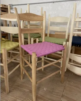 Several ladder back chairs are shown, some stacked on each other, in a variety of states of finish. There is a chair in the foreground with a pink wovven 