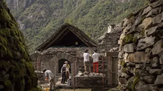In the foreground, old stone walls frame the sides of the photo. Between and behind them, people can be seen working on a stone house in a lush, mountainous region. There are forested hills visible in the far background.