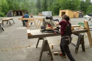 A student shot from the back is using a chop saw to cut lumber. There are other students working in the background.
