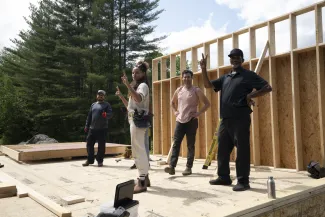 Four people are standing on a floor deck with one framed wall raised behind them. The four people are evenly spaced and facing the camera. Some are smiling and two are holding up peace signs.