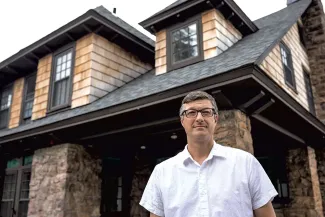 Devin Coleman stands in front of a cottage with stone and wood siding and gables. Devin is wearing a short sleeved white button down shirt, glasses, and short grey hair.