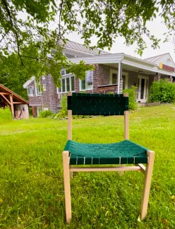 A maple chair with green woven seat and back is pictured outside in front of the main building on the lawn with tree branches overhead.