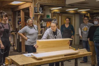 An instructor holds a piece of wood on a work bench, demonstrating for students standing behind.