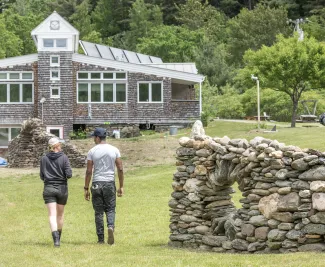 Two people walk towards the main building past a stone wall.