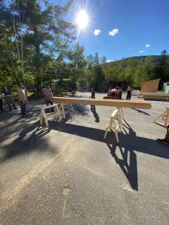 A group of people tests sawhorse strength by piling lumber on two sawhorses. It is a sunny day on blacktop with trees in the background.