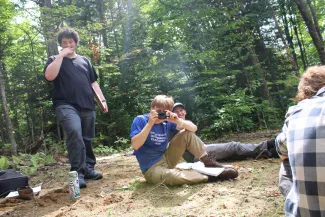 Three semester students are pictured in a sunny wooded clearing. The person in the center is pointing a camera at the photo taker, and there is another person standing to their left and one sitting to their right with their back turned.