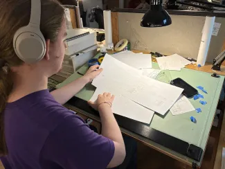 Photo looking over the shoulder of a student at a drafting table sifting through drawn plans. The student is wearing a purple t-shirt and silver headphones, with long brown hair pulled back in a low ponytail.