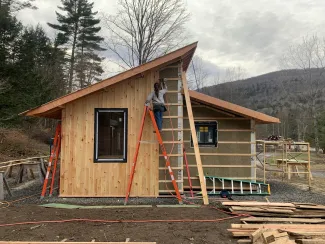 Sophia is pictured on a ladder working on the exterior siding of a small house. It is a gray day in the late fall.