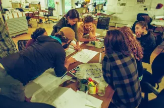 A group of students huddles around a table strewn with design drawings in the main studio