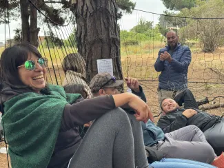 A person with dark hair and bangs, wearing a green shawl and sunglasses, smiles at the camera sitting in a tree net outside. There are other students/workers in the background.